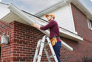 A worker standing on a ladder installs a white gutter along the roofline of a brick house.