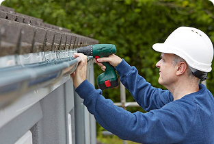 A construction worker wearing a white hard hat uses a cordless power drill to secure a metal railing along the edge of a structure